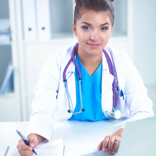 Schöne junge lächelnde Ärztin sitzt am Schreibtisch und schreibt. — Stockfoto