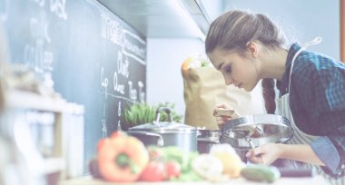 Genç bir kadın mutfağında yemek pişiriyor sobanın yanında duruyor.
