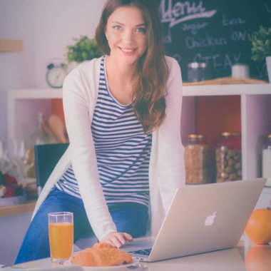 Dizüstü bilgisayar kullanarak ve mutfakta oturmuş çekici genç kadın