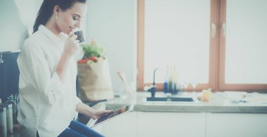 Genç bir kadın mutfakta tablet kullanıyor ve kahve içiyor.