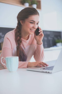 Genç kadın bir dizüstü bilgisayar kullanarak ve cep telefonuyla konuşurken mutfak masasında oturur