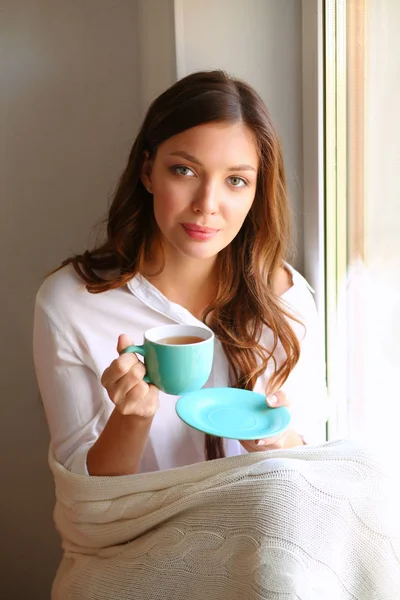 Młoda kobieta w domu siedzi w pobliżu okna relaksując się w salonie czytając książkę i pijąc kawę lub herbatę — Zdjęcie stockowe
