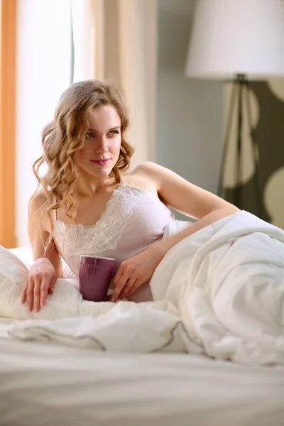 Νεαρή γυναίκα πίνοντας καφέ ή τσάι ξαπλωμένοι στο κρεβάτι — Φωτογραφία Αρχείου
