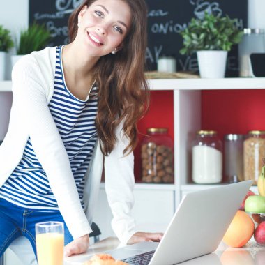 Dizüstü bilgisayar kullanarak ve mutfakta oturmuş çekici genç kadın