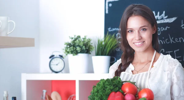 Mujer joven sonriente sosteniendo verduras de pie en la cocina —  Fotos de Stock
