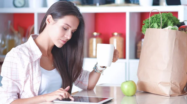 女人在她的厨房里喝茶的同时使用一台平板电脑 — 图库照片