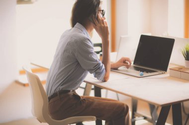 Güzel genç iş kadını ofis masasında oturuyor ve cep telefonuyla konuşuyor.