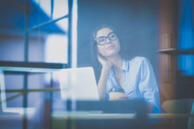 Ofis masasında dizüstü bilgisayarlı genç bir kadın oturuyor, pencereden bakıyor.