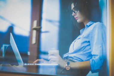 Ofis masasında dizüstü bilgisayarlı genç bir kadın oturuyor, pencereden bakıyor.