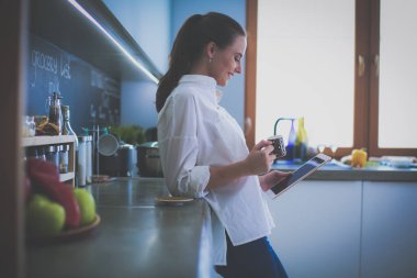 Genç bir kadın mutfakta tablet kullanıyor ve kahve içiyor.