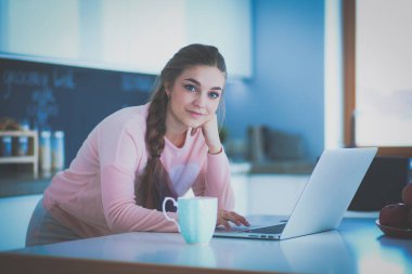 Genç kadın bir dizüstü bilgisayar kullanarak ve cep telefonuyla konuşurken mutfak masasında oturur