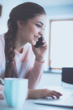 Genç kadın bir dizüstü bilgisayar kullanarak ve cep telefonuyla konuşurken mutfak masasında oturur