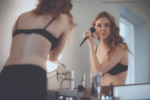 Молодая женщина смотрит в зеркало и наносит макияж — стоковое фото