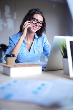 Güzel genç iş kadını ofis masasında oturuyor ve cep telefonuyla konuşuyor.