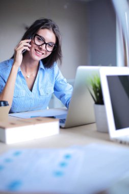Güzel genç iş kadını ofis masasında oturuyor ve cep telefonuyla konuşuyor.