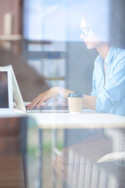 Ofis masasında dizüstü bilgisayarlı genç bir kadın oturuyor, pencereden bakıyor.