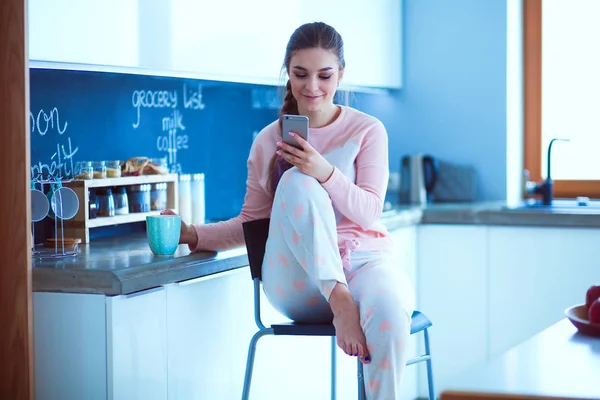 Mujer usando el teléfono móvil sentado en la cocina moderna — Foto de Stock