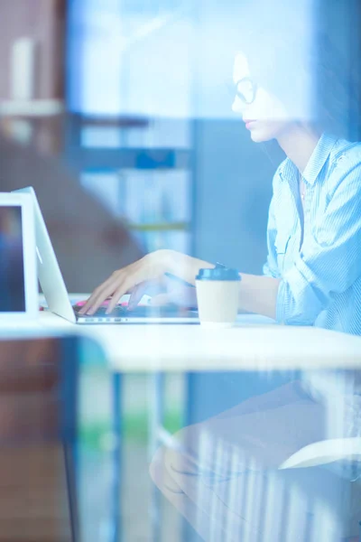 Jovem mulher sentada à mesa do escritório com laptop, vista através da janela — Fotografia de Stock