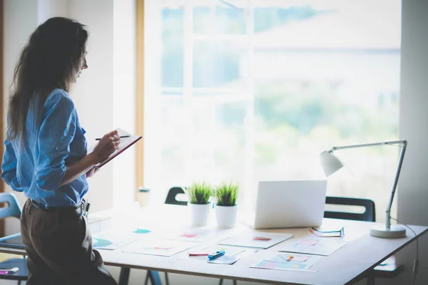 Jeune femme debout près du bureau avec des instruments, plan et ordinateur portable — Photo