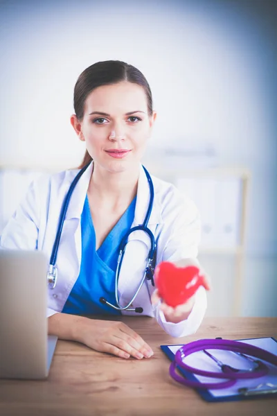 Schöne junge lächelnde Ärztin sitzt am Schreibtisch und hält Herz — Stockfoto