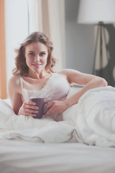 Mujer joven bebiendo taza de café o té mientras está acostada en la cama —  Fotos de Stock