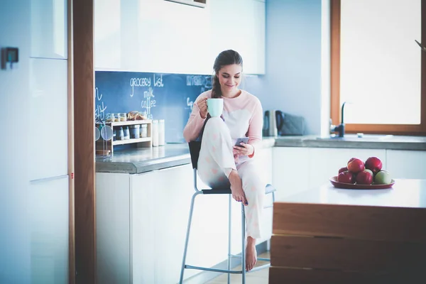 Mulher usando telefone celular sentado na cozinha moderna — Fotografia de Stock