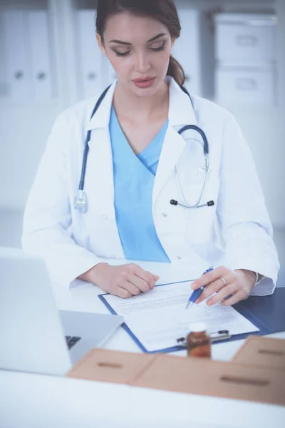 Schöne junge lächelnde Ärztin sitzt am Schreibtisch und schreibt — Stockfoto