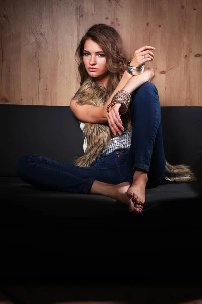 Porträt einer eleganten Frau, die auf einem schwarzen Sofa sitzt und eine blaue Jeans und Pelzweste trägt — Stockfoto