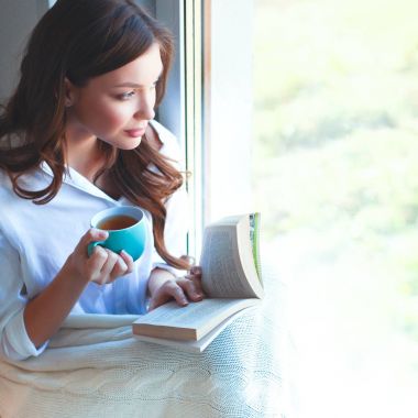 Evde oturan genç bir kadın oturma odasında oturmuş kitap okuyor ve çay ya da kahve içiyor.