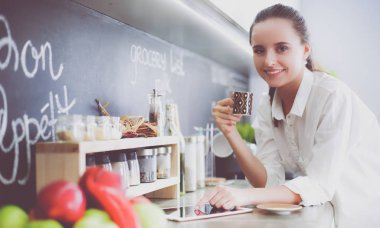 Genç bir kadın mutfakta tablet kullanıyor ve kahve içiyor.