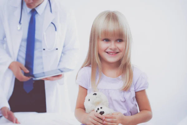 Little girl with teddy bear is looking at the camera. Female doctor on background — Stock Photo, Image