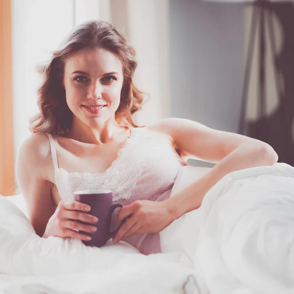 Mujer joven bebiendo taza de café o té mientras está acostada en la cama —  Fotos de Stock
