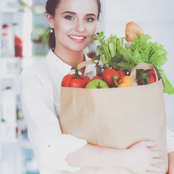 Mujer joven sosteniendo bolsa de la compra de comestibles con verduras. De pie en la cocina —  Fotos de Stock