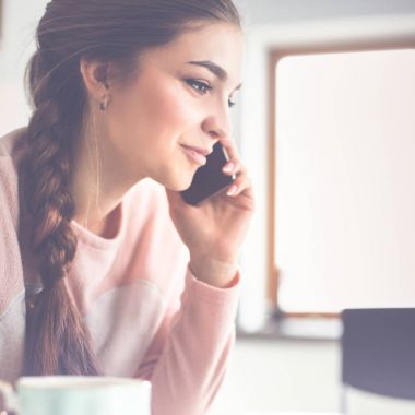 Genç kadın bir dizüstü bilgisayar kullanarak ve cep telefonuyla konuşurken mutfak masasında oturur