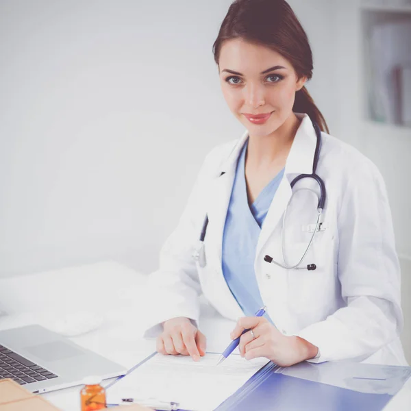 Όμορφη νεαρή χαμογελαστή γυναίκα γιατρός κάθεται στο γραφείο και γράφει — Φωτογραφία Αρχείου