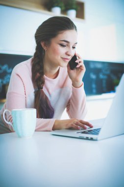 Genç kadın bir dizüstü bilgisayar kullanarak ve cep telefonuyla konuşurken mutfak masasında oturur.