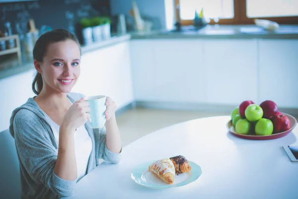 Kvinna som dricker te med söt croissant på köksbordet. — Stockfoto