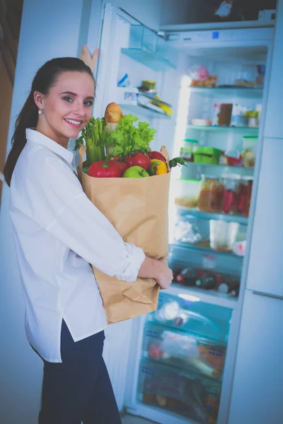 야채가 담긴 식료품 봉지를 들고 있는 젊은 여성이 부엌에 서 있습니다. 주방에서 카메라를 보고 있는 여자 — 스톡 사진