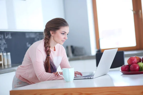 Mujer joven se sienta en la mesa de la cocina usando un ordenador portátil y hablando —  Fotos de Stock