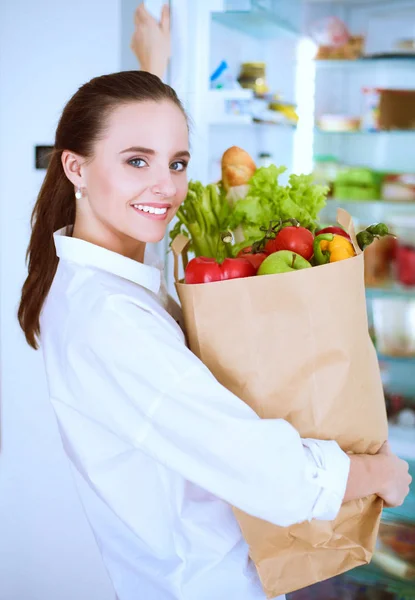 Mujer joven sosteniendo bolsa de la compra de comestibles con verduras.De pie en la cocina. Mujer en la cocina mirando a la cámara — Foto de Stock