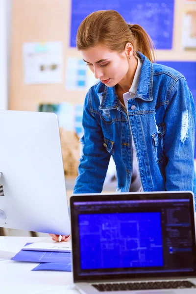 Fiatal nő áll íróasztal mellett eszközökkel, terv és laptop. — Stock Fotó