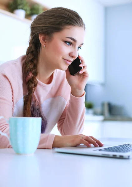 Junge Frau sitzt mit Laptop am Küchentisch und telefoniert. — Stockfoto