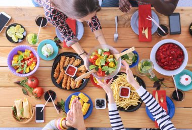 Ahşap masada otururken birlikte yemek yiyen bir grup insanın üst düzey görüntüsü. Masada yemek var. İnsanlar fast food yiyor..