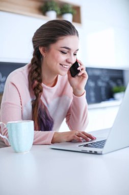 Genç kadın bir dizüstü bilgisayar kullanarak ve cep telefonuyla konuşurken mutfak masasında oturur.