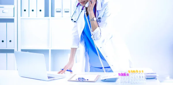 Молода жінка-лікар у білому пальто за комп'ютером за допомогою телефону — стокове фото