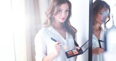 Genç ve güzel bir kadın aynanın yanında makyaj yapıyor.