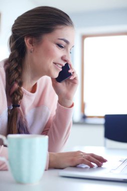 Genç kadın bir dizüstü bilgisayar kullanarak ve cep telefonuyla konuşurken mutfak masasında oturur.