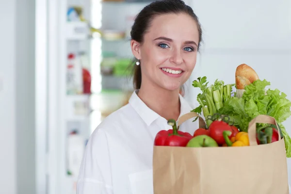 Ung kvinna som håller livsmedelsbutiker shoppingväska med grönsaker. Stående i köket. — Stockfoto