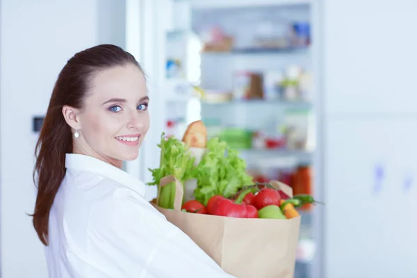 Молодая женщина держит продуктовый пакет с овощами. Женщина на кухне смотрит в камеру — стоковое фото