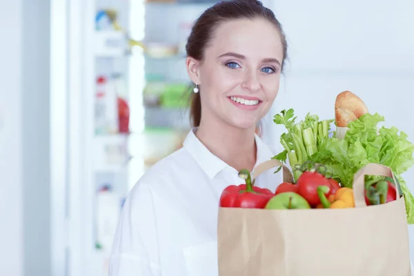 Ung kvinna som håller livsmedelsbutiker shoppingväska med grönsaker. Stående i köket. — Stockfoto